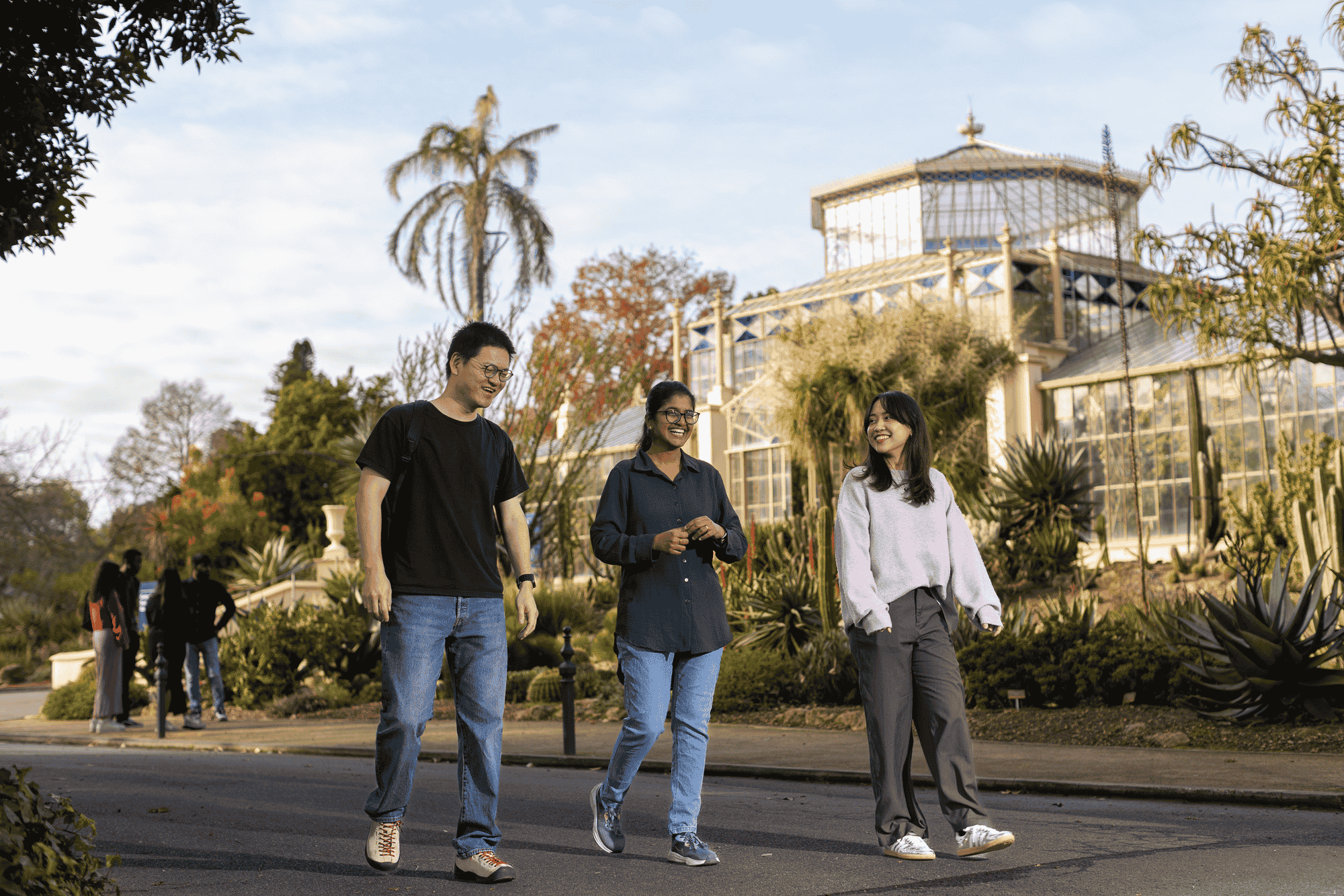 A group of five young adults walking outdoors on a sunny day, smiling and laughing together. They appear to be enjoying a casual outing in a park or market setting, with trees and people in the background.